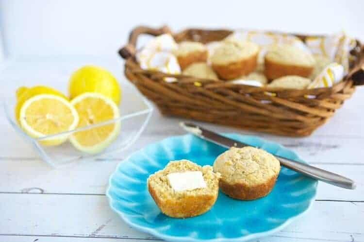 lemon ricotta gluten free muffins in a basket with one muffins sliced in half on a plate topped with butter. Also pictured is a butter knife and a bowl with lemons.