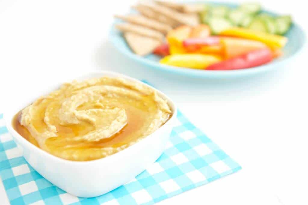 honey drizzled roasted garlic hummus in a bowl next to a plate of pita and veggies