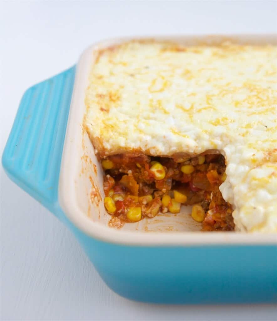 Taco Lasagna in a baking dish with one slice removed, showing the layers of corn, tomatoes, cheese, and tortillas. 