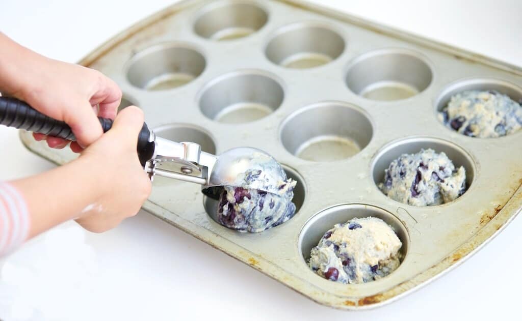using an ice cream scoop to fill muffin containers with blueberry muffin batter
