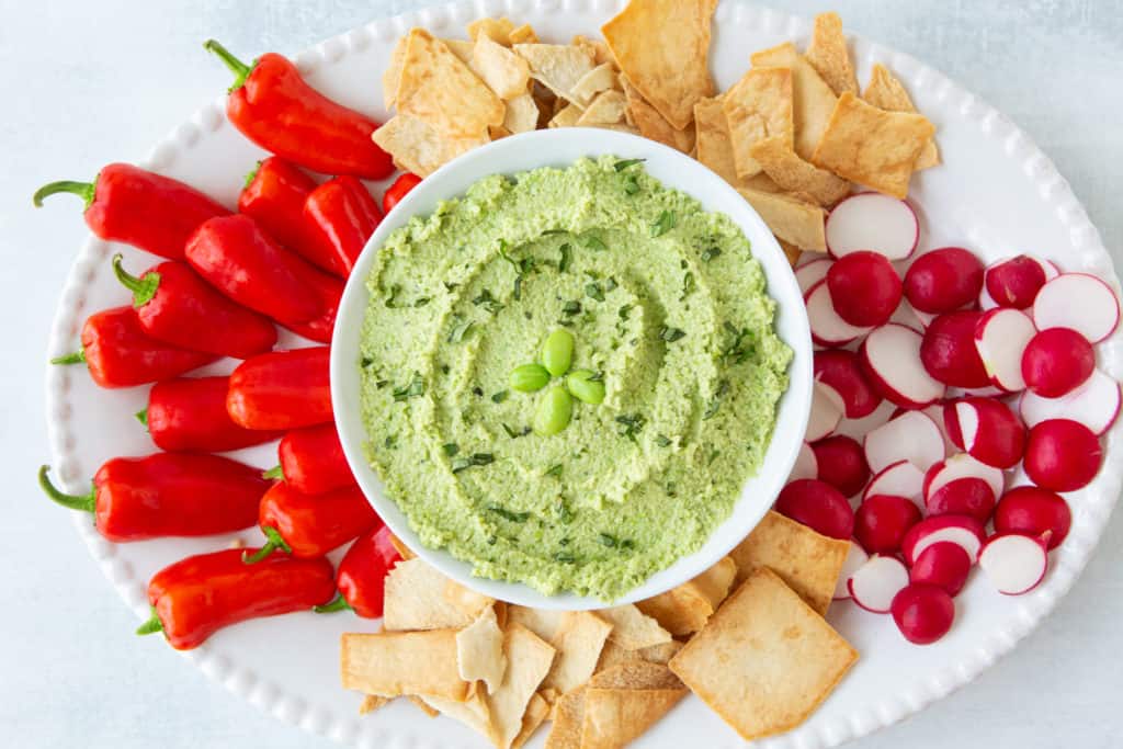 Green healthy hummus recipe with peppers, pita chips, and radishes