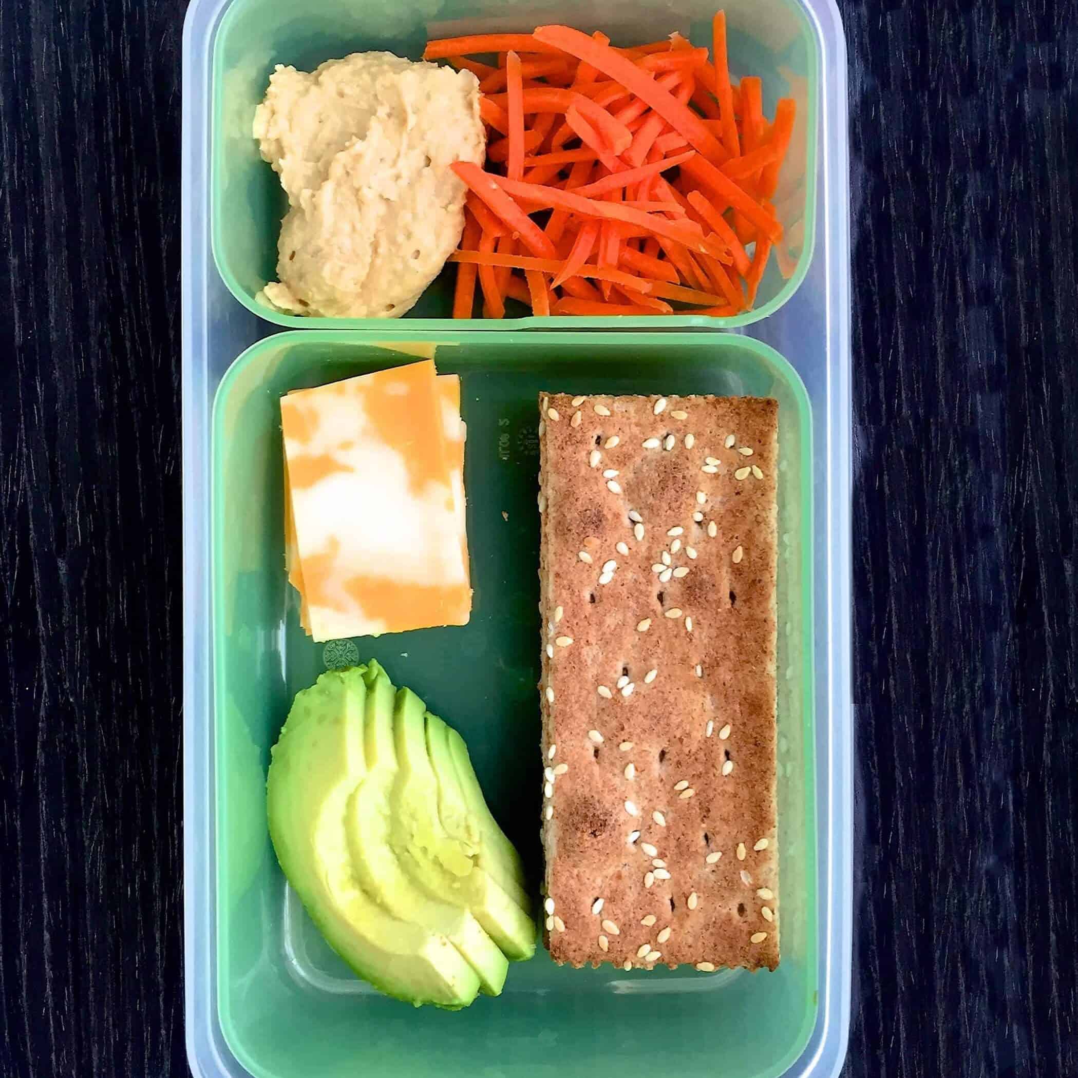 easy school lunch below contains whole grain crackers, cheese, hummus,  avocado slices, and shredded carrots